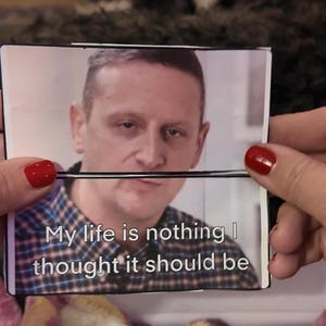 May include: A printed card held between two fingers with red nail varnish. The card features a man's portrait and the text: "My life is nothing I thought it should be."