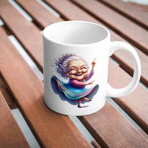 May include: White ceramic mug featuring a colourful illustration of a smiling woman with grey hair wearing a colourful skirt and glasses. She is dancing with her arms raised.