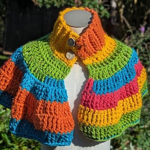 Puede incluir: Un chal de ganchillo colorido en un arcoíris de tonos: naranja, azul, verde, amarillo y rosa. El chal presenta un cuello alto y se abrocha con dos botones decorativos. El artículo hecho a mano se exhibe en un maniquí.
