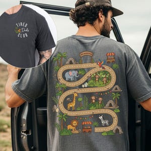 May include: Dark gray t-shirt with a jungle-themed graphic on the back featuring a road with animals like an elephant, giraffe, and zebra. The front left chest has the text "TIRED DADS CLUB" in white.