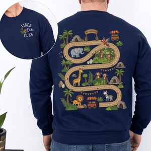 May include: Navy blue sweatshirt with a colorful safari scene on the back, featuring a road with animals like a giraffe, lion, and zebra. The front left chest has the text "Tired Dads Club".
