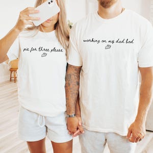 May include: Two matching white t-shirts with black text. One shirt says "pie for three please" and the other says "working on my dad bod". Both shirts have a small pie graphic. The people are wearing gray shorts.