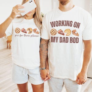 May include: Two people wearing matching white t-shirts and gray shorts are holding hands. The shirts have pie graphics and text. One shirt says "pie for three please" and the other says "WORKING ON MY DAD BOD".