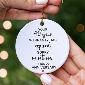 May include: A round white ceramic ornament featuring black text that reads: YOUR 40 year WARRANTY HAS expired SORRY no returns. HAPPY ANNIVERSARY with a small red heart icon at the bottom. The ornament hangs from a gold string.