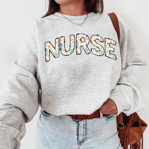 May include: A gray sweatshirt with the word "NURSE" printed in large, bold letters. The letters are filled with a floral pattern in shades of pink, yellow, and blue.