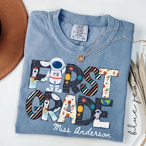May include: A blue short-sleeved t-shirt with a graphic print featuring the text FIRST GRADE and Miss Anderson. The design includes illustrations of astronauts, rockets, planets, and stars. The shirt has a Comfort Colors brand label.