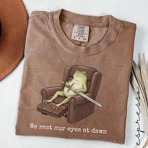 May include: Espresso-colored t-shirt featuring a relaxed frog in a brown recliner, holding a sword. The frog is light green with a white belly. The text "We rest our eyes at dawn" is printed at the bottom. The shirt has a Comfort Colors tag.