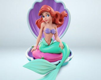 May include: A red-haired mermaid figurine sitting inside a purple seashell. The mermaid is wearing a purple bikini top and a green tail. The seashell is on a pink cushion.