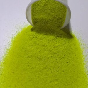 May include: A close-up of a bright green glitter, poured out of a container onto a white surface.