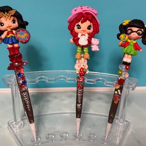 May include: Three decorative pens featuring character toppers. The left pen has a Wonder Woman figure, the center pen has a Strawberry Shortcake figure, and the right pen has a girl figure with pigtails. Each pen is adorned with colorful beads.