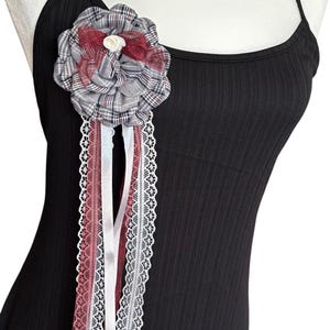 May include: A decorative flower pin with a plaid pattern in gray, white, and burgundy. The pin is adorned with burgundy and white ribbons, and a white lace trim. The pin is attached to a black ribbed tank top.