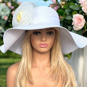 May include: A white sun hat with a wide brim and a decorative flower on the side. The hat features a textured weave and a ribbon band. The flower has a yellow center and white petals. The hat is designed for sun protection and fashion.