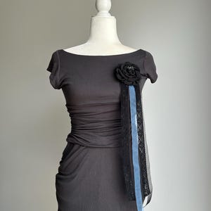 May include: Dark gray dress with a gathered waist and short sleeves. A black fabric flower with a blue ribbon and black lace detail is attached to the front. The dress is displayed on a white mannequin.