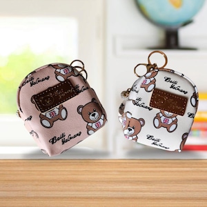 May include: Two small, white and pink, faux leather, backpack-shaped coin purses with gold-tone zipper pulls and keychains. The purses are decorated with a repeating pattern of brown teddy bears wearing red, white, and blue clothing. The text "Baili YaGuang" is printed on the purses.