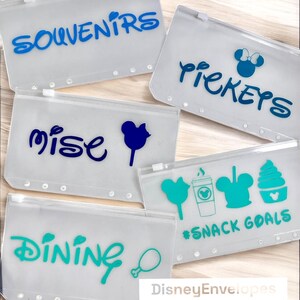 May include: Five translucent plastic zipper pouches with six-hole punched edges. The pouches feature blue, dark blue, and teal vinyl decals including the text SOUVENIRS, TICKETS, MISE, DINING, and #SNACK GOALS, alongside icons of mouse ears and food.