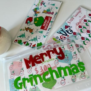 May include: Clear plastic budget envelopes with Grinch-themed designs. One envelope reads "Merry Grinchmas" with a $300 savings challenge. Other envelopes feature Grinch characters and savings challenges.