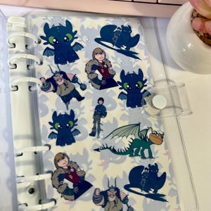 May include: A clear, plastic binder with a variety of colorful cartoon character stickers. The stickers feature characters from the "How to Train Your Dragon" franchise, including dragons and Viking characters. The binder is open, revealing the stickers inside.