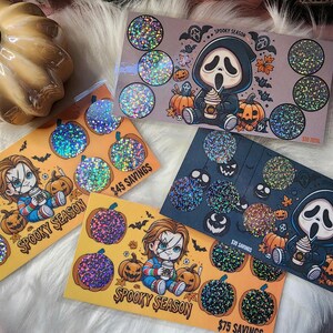 May include: Four holographic sticker sheets featuring a spooky season theme. Each sheet has a different character, including Ghostface, a character from the movie Scream, and a character from the movie Child's Play. The stickers are decorated with pumpkins, bats, and other Halloween imagery. The text on the stickers reads "Spooky Season" and includes the price savings for each sheet.