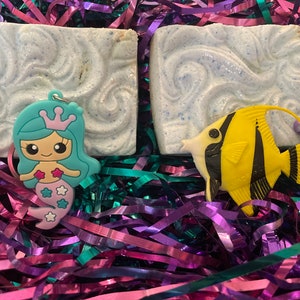 May include: Two square, light blue soaps with a wave design, a teal mermaid keychain, and a yellow and black fish-shaped charm rest on a bed of purple, teal, and pink shredded paper. The mermaid has a pink tail and a crown.