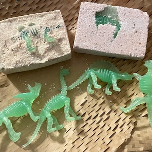 May include: Four green plastic dinosaur skeletons on a brown textured surface. Two of the skeletons are partially embedded in a white block of bath salts.