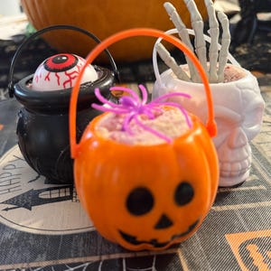 May include: Three small plastic Halloween buckets with handles. One is an orange pumpkin with a purple spider, one is a black cauldron containing a realistic eyeball, and one is a white skull filled with a grey skeletal hand reaching upward.