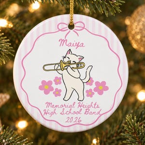 Puede incluir: Un adorno redondo de cerámica blanca con borde de rayas rosas y cordón dorado. Presenta un gato blanco de dibujos animados tocando un trombón dorado, flores rosas y el texto: Maiya, Memorial Heights High School Band y 2026.