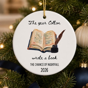Op de afbeelding: Een rond wit keramieken ornament dat aan een gouden koord hangt. Het midden toont een illustratie van een open boek met een ganzenveer en inktpot. De tekst luidt: The year Colton wrote a book, THE CHANCE OF NIGHTFALL, en het jaar 2026.