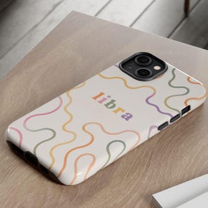 May include: A white phone case featuring a repeating pattern of wavy, multicolored lines in shades of orange, green, and purple. The word "libra" is printed in the center in a multicolored, sans-serif font, with a black camera cutout.