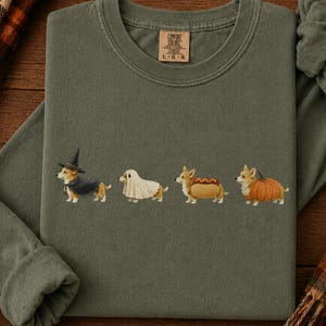 May include: An olive green crewneck sweatshirt featuring a graphic of four Corgi dogs in Halloween costumes: a witch, a ghost, a hot dog, and a pumpkin. The garment has a ribbed collar and a Comfort Colors brand label printed on the inner neck area.