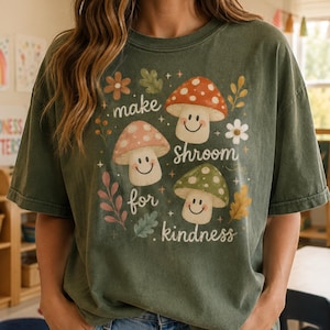 May include: A moss green short-sleeve t-shirt featuring a graphic print of three smiling mushrooms in red, tan, and green. The design includes small flowers, leaves, and stars with the text "make shroom for kindness" written in a cursive white font.