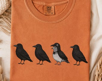 Może przedstawiać: T-shirt w kolorze wypalonej pomarańczy z nadrukiem czterech ptaków w rzędzie. Trzy ptaki to czarne wrony, a trzeci od lewej to szaro-czarny gołąb w czarnej obroży. Koszulka posiada okrągły dekolt i metkę marki Comfort Colors przy szyi.