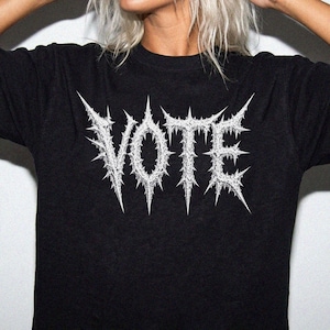 May include: A black short-sleeved t-shirt featuring the word VOTE printed in a white, jagged, thorn-like font across the chest. The design has a sharp, spiky aesthetic with intricate details within the lettering, set against a solid black fabric.