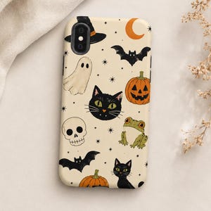 May include: A cream-colored phone case featuring a repeating pattern of Halloween-themed illustrations including black cats, orange pumpkins, a white ghost, a skull, a green frog, bats, a witch hat, and a crescent moon, accented with small black stars.
