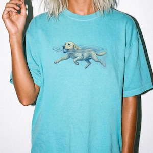 May include: A lagoon blue t-shirt featuring a graphic illustration of a light-colored dog swimming in water while holding a small blue ball in its mouth. The dog is depicted with ripples in the water surrounding it, centered on the chest area.
