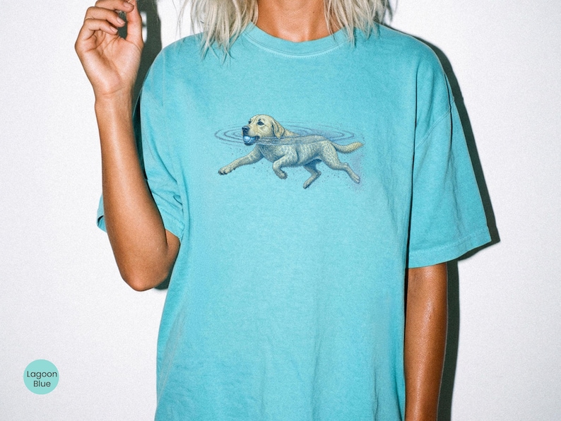 May include: A lagoon blue t-shirt featuring a graphic illustration of a light-colored dog swimming in water while holding a small blue ball in its mouth. The dog is depicted with ripples in the water surrounding it, centered on the chest area.