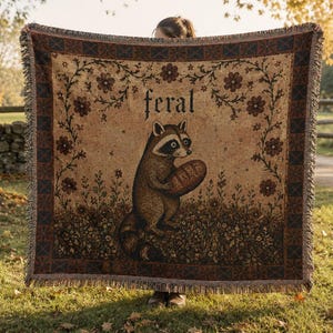 May include: A woven throw blanket featuring a central illustration of a raccoon holding a loaf of bread, with the word feral printed above. The design includes a floral border, earthy brown and tan tones, and a fringed edge on all four sides.