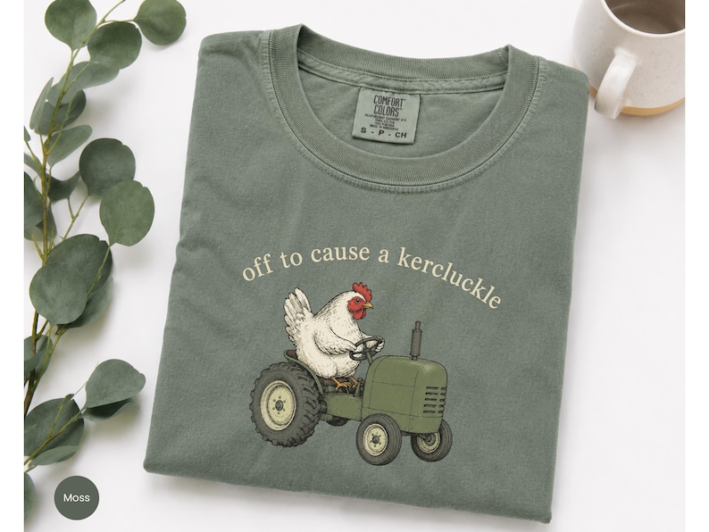 May include: A moss green t-shirt featuring a graphic of a white chicken driving a green tractor. The text above the chicken reads, off to cause a kercluckle. The shirt has a Comfort Colors brand label at the inner neckline showing size S-P-CH.