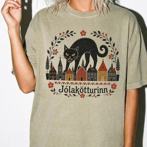 May include: A khaki-colored t-shirt featuring a graphic print of a black cat standing over a row of small houses. The design includes a floral wreath border, red and orange accents, and the text J&oacute;lak&ouml;tturinn printed in a dark serif font.