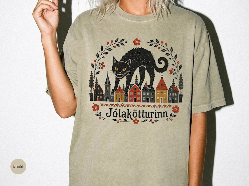 May include: A khaki-colored t-shirt featuring a graphic print of a black cat standing over a row of small houses. The design includes a floral wreath border, red and orange accents, and the text J&oacute;lak&ouml;tturinn printed in a dark serif font.