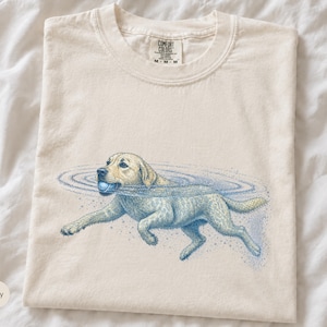 May include: An ivory-colored crew neck t-shirt featuring a graphic illustration of a yellow Labrador retriever swimming in water while holding a blue ball in its mouth. The shirt has a Comfort Colors brand label printed on the inside neck area.