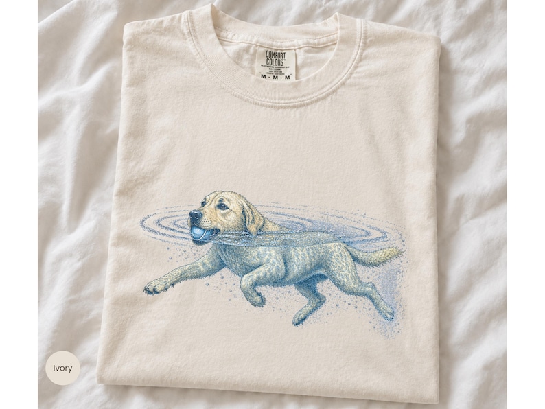 May include: An ivory-colored crew neck t-shirt featuring a graphic illustration of a yellow Labrador retriever swimming in water while holding a blue ball in its mouth. The shirt has a Comfort Colors brand label printed on the inside neck area.