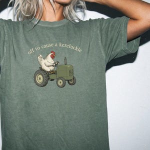 May include: A moss green short-sleeve t-shirt featuring a graphic of a white chicken driving a green tractor. Above the illustration, the text reads "off to cause a kercluckle" in a simple, light-colored, lowercase font.