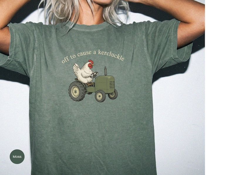 May include: A moss green short-sleeve t-shirt featuring a graphic of a white chicken driving a green tractor. Above the illustration, the text reads "off to cause a kercluckle" in a simple, light-colored, lowercase font.