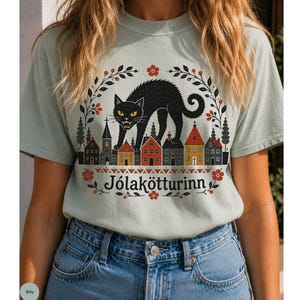 May include: A light sage green t-shirt featuring a graphic of a black cat arching its back over a row of small houses and a church. The design includes a floral wreath border and the text J&oacute;lak&ouml;tturinn printed in black below the illustration.
