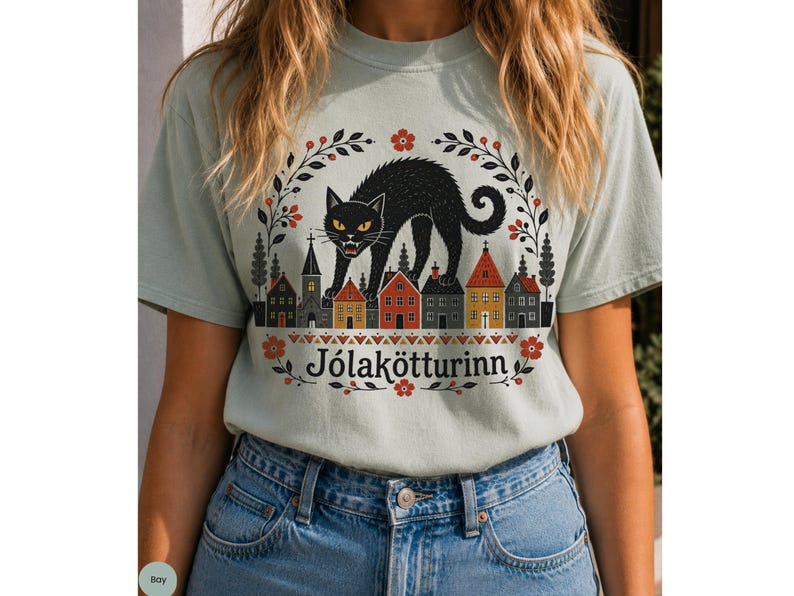 May include: A light sage green t-shirt featuring a graphic of a black cat arching its back over a row of small houses and a church. The design includes a floral wreath border and the text J&oacute;lak&ouml;tturinn printed in black below the illustration.