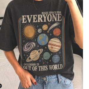 May include: A black short-sleeved t-shirt featuring a graphic print of the solar system with planets, stars, and a galaxy. The text reads "MAKE SPACE FOR EVERYONE" at the top and "INCLUSION IS OUT OF THIS WORLD" at the bottom in a retro font.