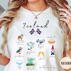 Könnte beinhalten: Cremefarbenes T-Shirt mit dem Wort "Iceland" und Illustrationen von Island-Elementen, wie Islandpferden, Papageientauchern und Nordlichtern. Das T-Shirt zeigt auch die isländische Flagge und das Wort "NATURAL".