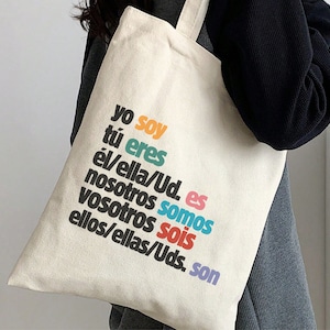 Op de afbeelding: Een witte canvas draagtas met de Spaanse vervoeging van het werkwoord "zijn" bedrukt in kleurrijke letters. De tekst luidt: "yo soy, tú eres, él/ella/Ud. es, nosotros somos, vosotros sois, ellos/ellas/Uds. son".