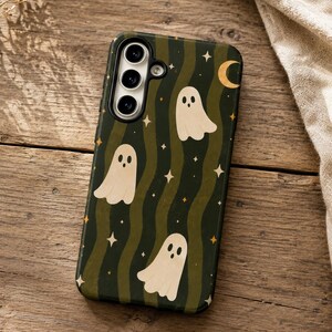 May include: A rectangular phone case featuring a dark green and olive wavy striped background with a pattern of cream-colored ghosts, small four-pointed stars, and a crescent moon. The case has a cutout for a triple-lens camera system.