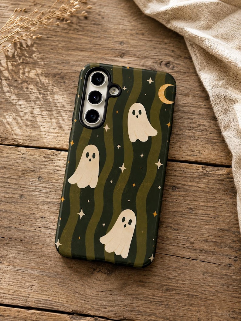 May include: A rectangular phone case featuring a dark green and olive wavy striped background with a pattern of cream-colored ghosts, small four-pointed stars, and a crescent moon. The case has a cutout for a triple-lens camera system.